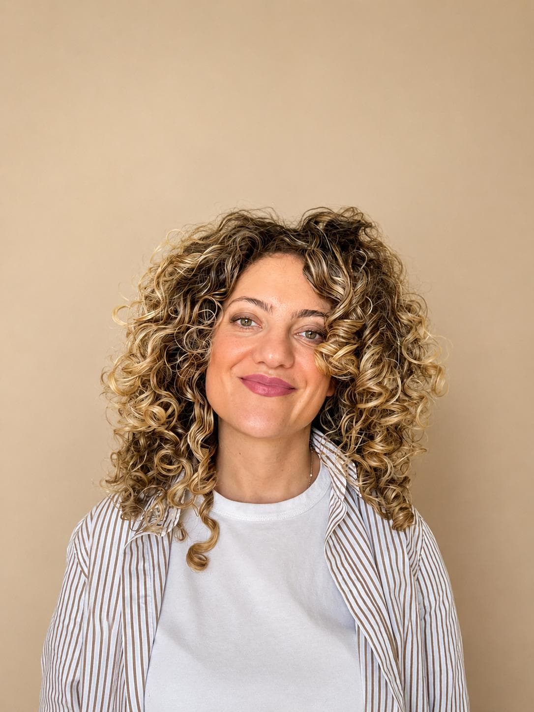 Mulher sorridente com volumosos cabelos cacheados loiros, vestindo camiseta branca e camisa listrada aberta.